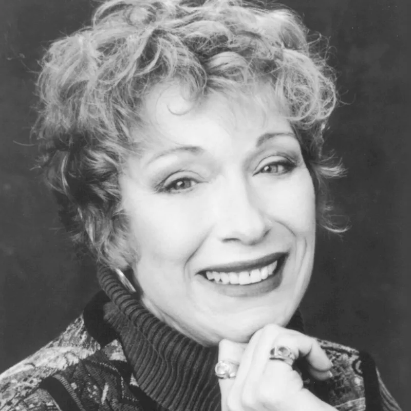 Black-and-white head-and-shoulders portrait of a smiling woman with short curly hair, resting her chin on her hand and wearing a dark turtleneck and patterned jacket.