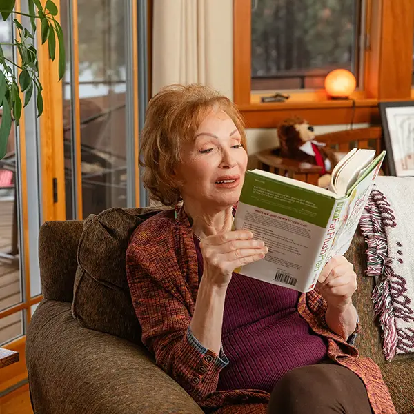 Older woman relaxing on a sofa while reading a green-covered cookbook in a cozy living room with warm lighting and a knit throw nearby.